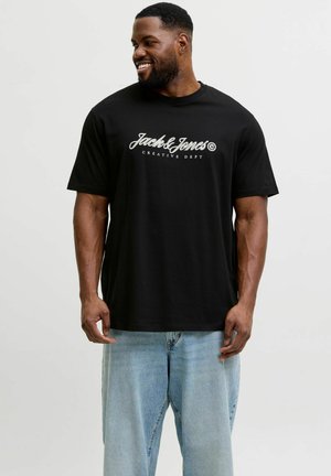 Mosolygó férfi fekete pólóban, amelyen a „Jack & Jones Creative Dept” felirat olvasható, világoskék, bő szabású farmerrel, fehér háttér előtt áll.