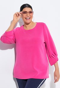 Top rosa oversize a maniche lunghe con scollatura rotonda e dettagli arricciati sulle maniche, abbinato a pantaloni blu scuro e occhiali grandi e trasparenti.