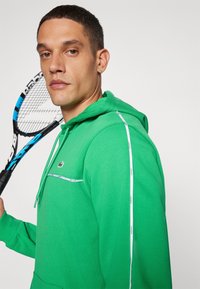 Sweatshirt verde com bolso canguru, apresentando riscas laterais brancas e um pequeno logótipo da Lacoste. Feito de um tecido macio e texturizado.