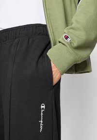 Veste à fermeture éclair vert olive avec poignets côtelés, associée à un pantalon de survêtement noir avec poches latérales et logo blanc brodé.