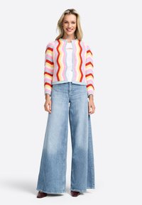 Cardigan tricot coloré avec un motif en zigzag rose, rouge et jaune, associé à un jean en denim bleu clair à jambe large et des talons bordeaux.