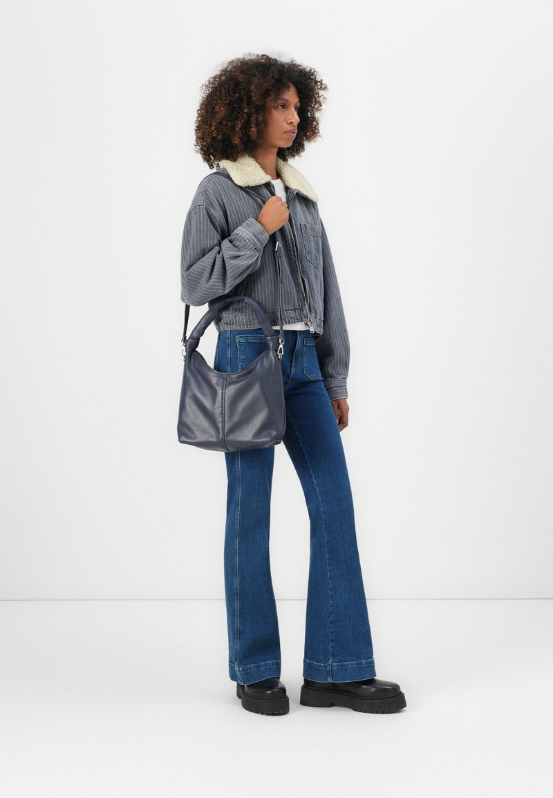 Dunkelgraue Cordjacke mit einem Schaffellkragen, weit geschnittene blaue Jeans, schwarze klobige Stiefel und eine marineblaue Ledertasche in runder Form.