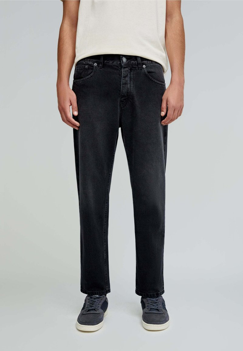 Jeans negre din denim cu tăietură dreaptă, cinci buzunare și detalii de cusătură subtile, asortați cu adidași gri. Design simplu și casual.