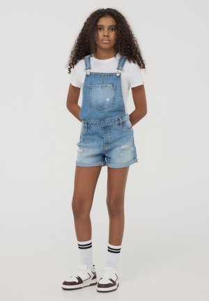 Pantaloni salopette in denim azzurro chiaro, con tasca, pantaloni corti con orlo ripiegato, dettagli strappati e spalline regolabili, indossati sopra una maglietta bianca.