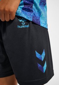 Svarta träningsbyxor med blå "hummel"-logotyp och chevronmönster. Mjuk tyg med fukttransporterande textur, lämplig för sportkläder.