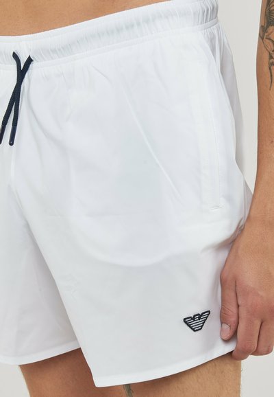 Gros plan d'une personne portant un short de bain blanc avec des cordons noirs et un petit logo noir sur la jambe gauche près de l'ourlet.
