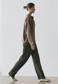 Vrouw loopt naar rechts en draagt een loszittend bruin shirt met lange mouwen, donkere wijd uitlopende broek en lichtgekleurde ballerina's tegen een effen achtergrond.