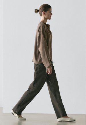 Eine Frau geht nach rechts, trägt ein weites braunes Langarmshirt, dunkle weit geschnittene Hosen und helle Ballerinas vor einem schlichten Hintergrund.