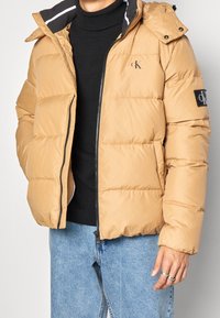 Beige pufferjakke med lynlås fortil, elastiske ærmer, aftagelig hætte og logo-patch på ærmet. Båret over en sort trøje og jeans.