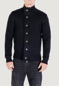 Uomo che indossa un maglione a maglia nero con bottoni e un colletto alto, insieme a jeans neri lavati, in piedi di fronte a uno sfondo semplice.