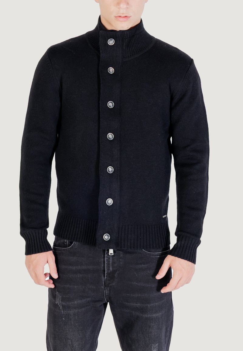 Uomo che indossa un maglione a maglia nero con bottoni e un colletto alto, insieme a jeans neri lavati, in piedi di fronte a uno sfondo semplice.