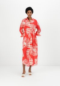 Rochie roșie cu butoni, cu un model floral alb în spirală, mâneci lungi, talie strânsă și un design până la genunchi, asociată cu sandale de culoare deschisă.