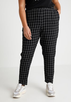 Person trägt schwarze Hose mit weißem Gitternetzmuster, weiße Sneakers und ein schwarzes Oberteil, steht mit einer Hand in der Tasche.
