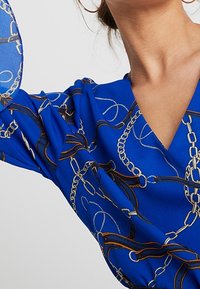 Modré šaty s výstřihem do V, s potiskem řetězce a pásku ve zlaté a stříbrné barvě. Hladká tkanina, krátké rukávy a volný střih.