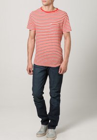 Camiseta de manga corta a rayas rojas y blancas con cuello redondo y bolsillo en el pecho, combinada con jeans de mezclilla azul oscuro y zapatillas de color claro.