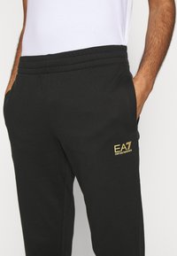 Svarta sweatpants i bomullsblandning med elastisk midja, sidofickor och en guldfärgad logotyp på vänster lår.