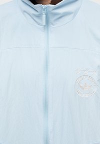 Chaqueta ligera de color azul claro con cremallera completa y capucha. Presenta un logotipo circular bordado con tres estrellas en el pecho.