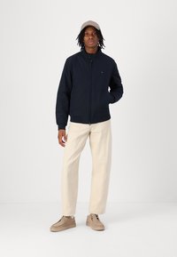 Veste zippée marine avec col haut, casquette beige, pantalon crème et chaussures beige clair. Design simple, texture lisse et couleurs contrastantes.