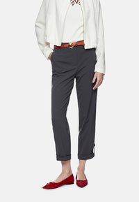 Graue, maßgeschneiderte Hosen mit hochgekrempelten Säumen, kombiniert mit einer weißen Bomberjacke. Ergänzt durch einen roten Gürtel und rote, mit einer Schleife verzierte Ballerinas.