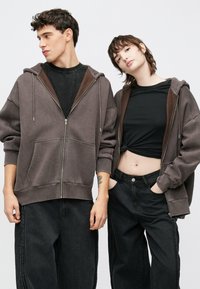 Δύο μοντέλα στέκονται δίπλα δίπλα φορώντας φαρδιές, φούτερ με φερμουάρ σε σκούρο καφέ και μαύρο. Τα φούτερ έχουν μεγάλες τσέπες και ριμπ μανσέτες.