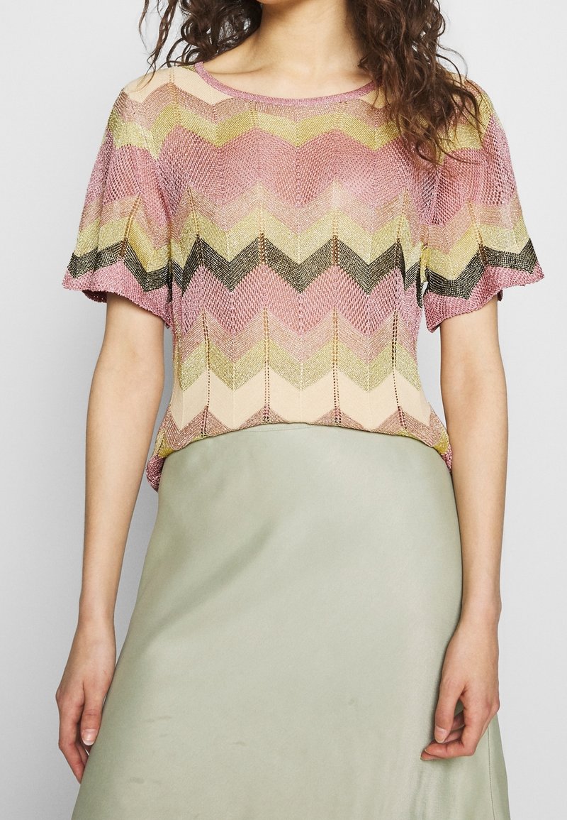 Knit top featuring a chevron pattern in pink, yellow, and black tones. Short sleeves and a round neckline, paired with a smooth, green skirt.