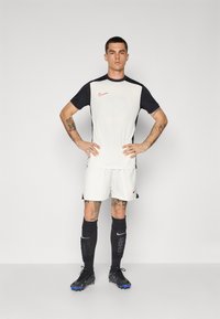 Biała koszulka sportowa z czarnymi rękawami, z czerwonym logo Nike. Białe spodenki i czarne skarpety. Czarne korki piłkarskie z niebieskimi akcentami.