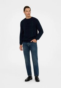 Temno modra pletena majica z vzorcem kit, nošena s temnimi jeans hlačami in črnimi čevlji. Tkanina deluje mehko z vidnim teksturiranim zaključkom.