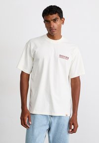 T-shirt bianco in cotone con collo rotondo, caratterizzato da un piccolo logo rosso sul petto sinistro. Abbinato a jeans in denim blu chiaro.
