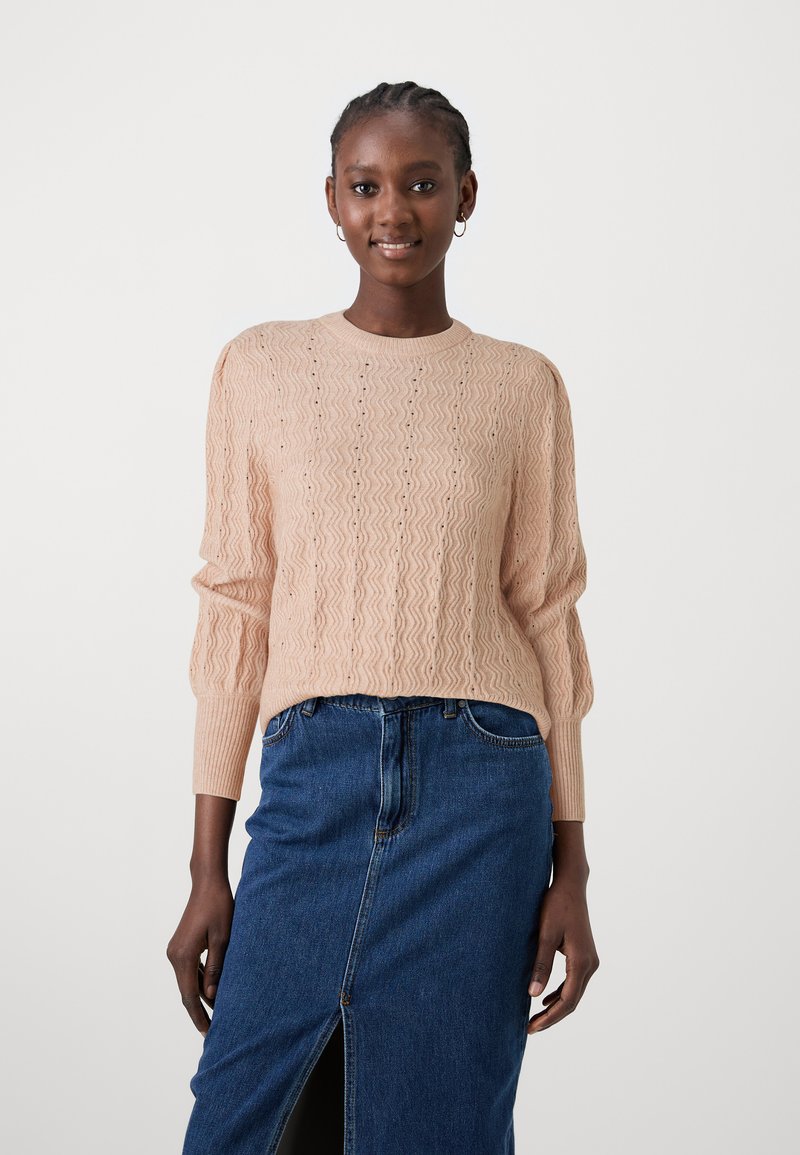 Pull en tricot rose clair avec un motif ondulé, à manches longues et avec un ourlet côtelé. Associé à une jupe en denim foncé avec une fente à l'avant.