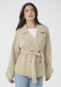 Beige trench coat dengan desain double-breasted, pinggang berikat, epaulet bahu, dan kancing besar. Kain bertekstur dan lengan dilipat.