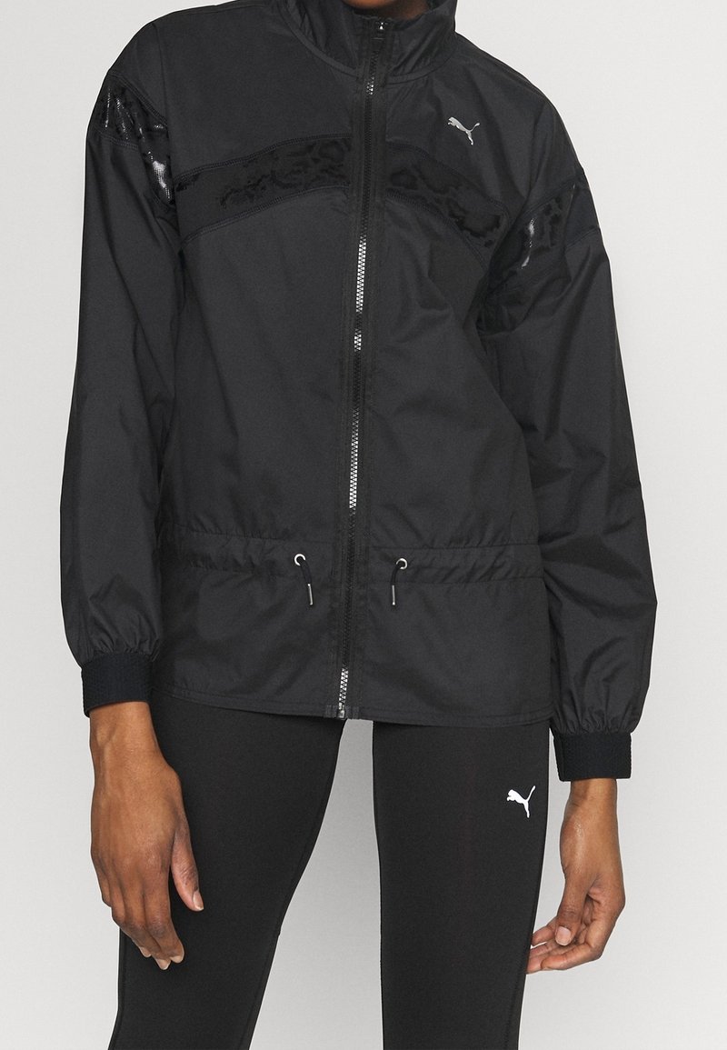 Schwarze Windjacke mit Reißverschluss vorne, elastischen Bündchen und Kordelzugsaum. Verfügt über ein glänzendes Panel und ein Logo auf der Brust. Glatte Textur.