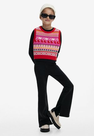 Bunter, gekürzter Pullover in Rot, Pink und Schwarz mit geometrischen Mustern. Schwarze Schlaghosen und beiges Schuhwerk mit schwarzen Sohlen. Weißer Beanie und Sonnenbrille.