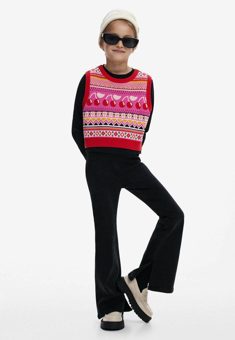 Maglione corto colorato in rosso, rosa e nero con motivi geometrici. Pantaloni neri a campana e scarpe beige con suole nere. Berretto bianco e occhiali da sole.