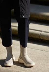 Beige Knöchelstiefel mit dickem Gummisohle, glatter Lederstruktur und seitlichen elastischen Einsätzen, getragen mit dunklen, kurzen Hosen.