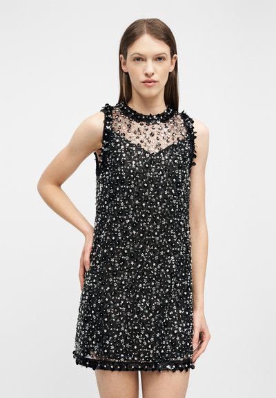 Femme portant une mini robe noire sans manches avec un panneau supérieur en tissu transparent et des embellissements floraux à paillettes superposés, debout devant un fond uni.