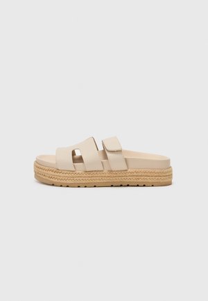 Vaaleat liukusandaalit, joissa on kaksi leveää remmiä, teksturoitu espadrille-tyylinen pohja, jossa on punottu juuttikoriste. Litteä, rento muotoilu.