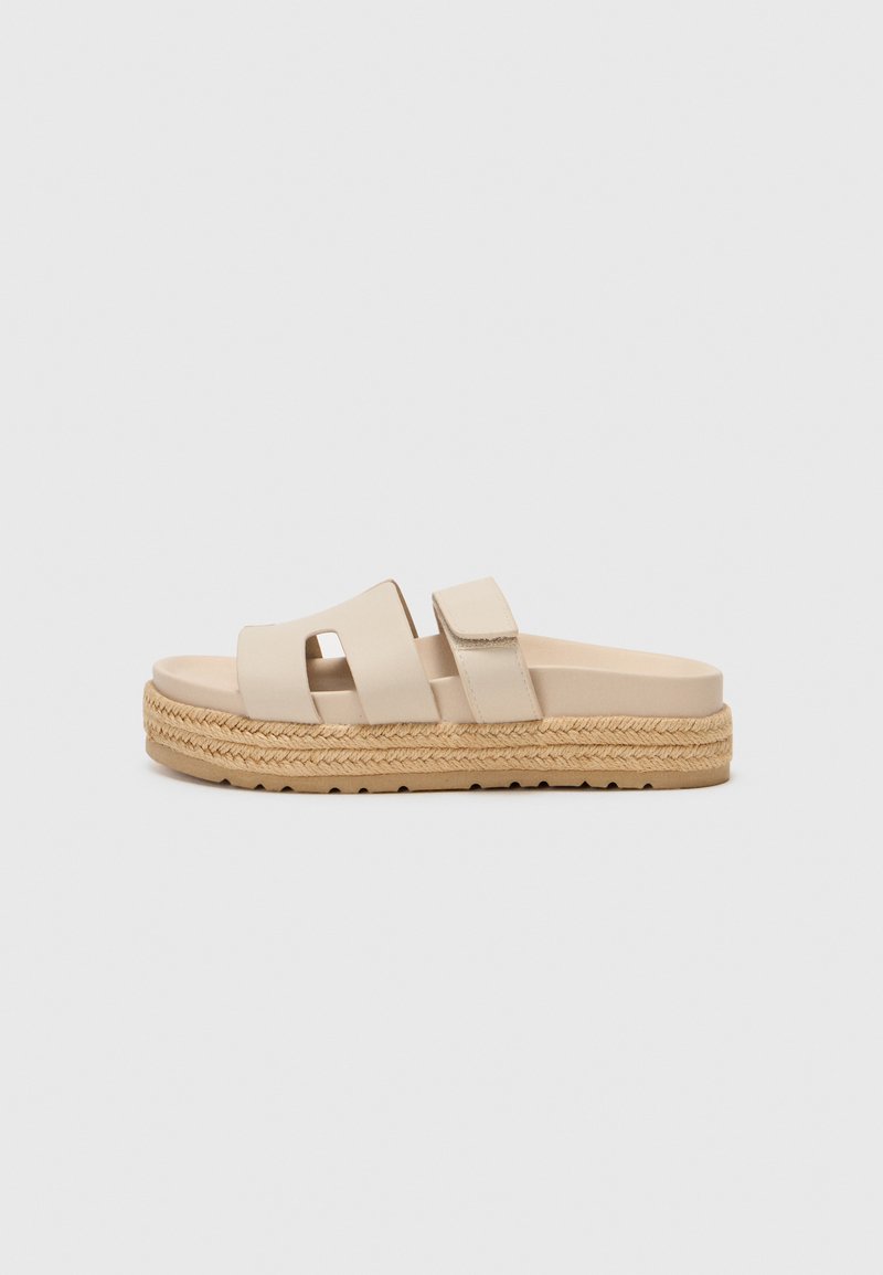 Beige slide sandals with two wide straps, a textured espadrille-style platform sole featuring a braided jute accent. Flat, casual design.
