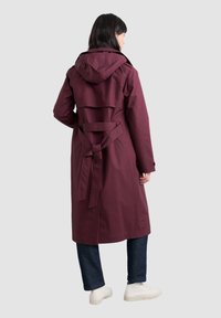 Bordeaux lang jasje met capuchon, tailleband en uitlopende rugdetail. Gekocht met donkere jeans en witte sneakers. Gladde stoftextuur.