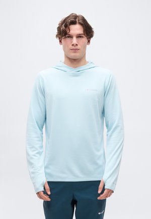 Mladi moški z valovitimi lasmi, oblečen v svetlo modro kapucarico in temno modre hlače, stoji pred enostavnim belim ozadjem.