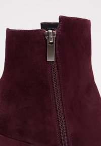 Bottines en daim bordeaux à fermeture éclair sur le côté, avec une texture lisse et des lignes épurées. Coutures d'accent visibles le long du bord.