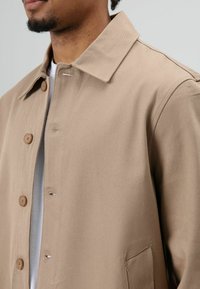 Beige katoenen jas met een kraag, knoopsluiting aan de voorkant en twee zijkanten zakken. Voorzien van een textuurstof en zichtbare stikdetails.