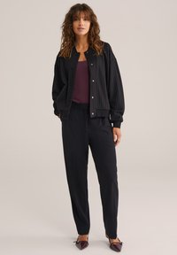 Juodas striukė su smulkia juostele virš burgundiško marškinėlio, derinama su juodomis kelnėmis ir smailiais bateliais, sukuria elegantišką, sluoksniuotą įvaizdį.