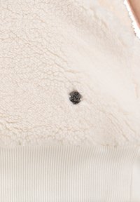 Tela de forro de color crema con una superficie texturizada y esponjosa, y un ribete blanco acanalado. Presenta un pequeño botón decorativo negro con texto.