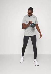 Muž v šedej košeli a čiernych legínach vykonáva cvičenie s kettlebellom, pričom sa pozerá na váhu.