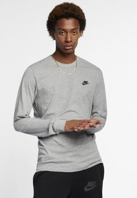 T-shirt de manga longa cinza da Nike com um pequeno logotipo preto no peito, combinado com calças pretas, com as mãos posicionadas à frente.