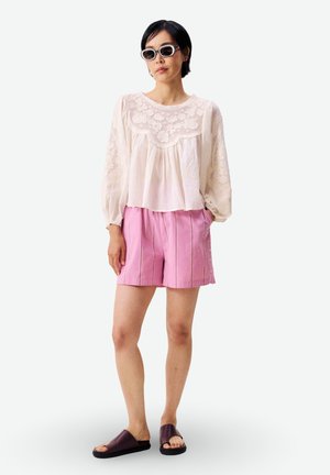 Femme portant un chemisier en dentelle blanche, un short plissé rose, des sandales plates foncées et des lunettes de soleil, debout avec une main dans la poche sur fond blanc.