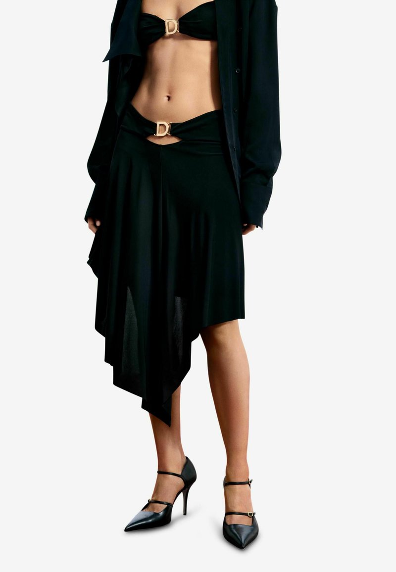 Falda asimétrica negra con drapeado, con una hebilla dorada en forma de D. Combinada con un top a juego sin tirantes y tacones negros puntiagudos.