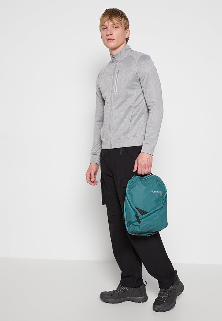 Mann steht in einer grauen Zip-up-Jacke, schwarzen Hosen und schwarzen Wanderschuhen, hält einen türkisfarbenen Rucksack an der Seite vor einem schlichten weißen Hintergrund.