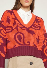 Femme portant un pull court en tricot à motif orange et bordeaux par-dessus une chemise blanche à col et un pantalon beige taille haute avec un bouton noir.