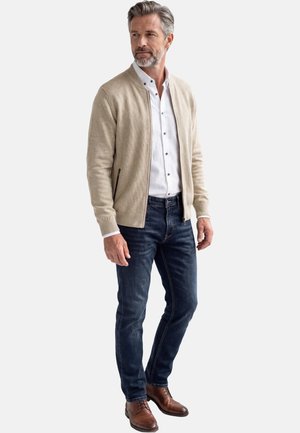 Middelbare man staand, gekleed in een beige vest met ritssluiting, wit overhemd, donkerblauwe spijkerbroek en bruine leren schoenen, licht naar links kijkend.